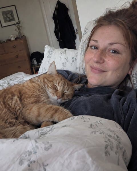 Chloe in bed with her ginger cat sleeping against her shoulder.